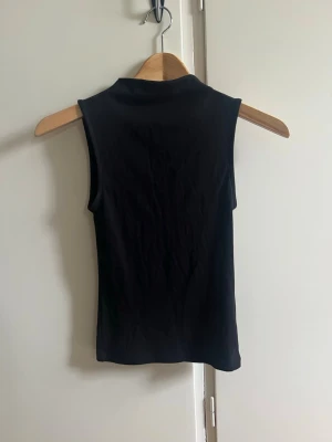 Svart tajt topp med korta ärmar och hög krage - Säljer en svart, ärmlös polotopp i mjukt material. Toppen har en hög krage och en slim passform som sitter snyggt på kroppen. Perfekt att styla med jeans eller kjol för en trendig look. Endast använd 1 gång. Välj gärna instabox