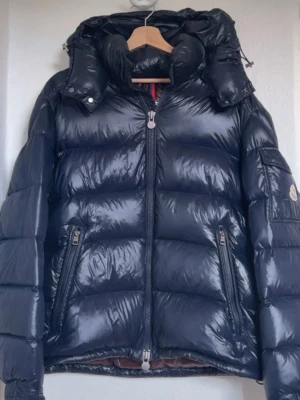 Moncler Maya jacka  - Snygg svart dunjacka från Moncler med glansig finish och avtagbar huva. Jackan har två dragkedjeförsedda fickor framtill och klassisk Moncler-logga på ärmen. Insidan har ett brunt foder med serietidningsmönster. Perfekt för kalla vinterdagar.