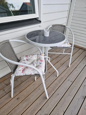 Runt cafébord med två stolar - Säljer ett runt cafébord med grå glasskiva och två matchande stolar i vitt och grått. Stolarna har flätad sits och rygg samt mjuka sittdynor med blommigt mönster. Perfekt för balkongen eller uteplatsen.