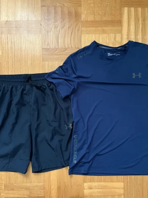 Träningsset från Under Armour - Snyggt träningsset från Under Armour med en marinblå t-shirt och svarta shorts. T-shirten har rund hals och diskret logga på bröstet, medan shortsen har elastisk midja och logga på benet. Perfekt för gymmet eller löpning.