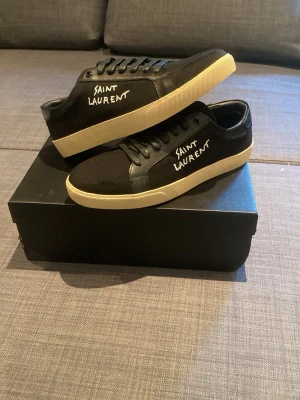 Svarta Saint Laurent sneakers - Säljer ett par svarta sneakers från Saint Laurent med vit sula och handskriven logga på sidan. Skorna har snörning och är tillverkade i en mix av skinn och textil. Klassisk låg modell som passar till det mesta. Är storlek 42 men sitter som 43