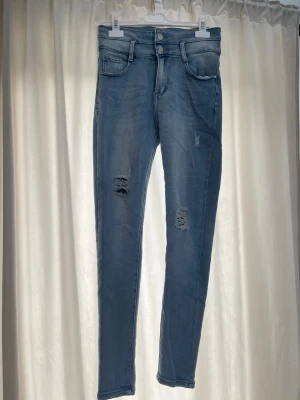 Ljusblå skinny jeans med slitningar - Säljer ett par ljusblå skinny jeans med trendiga slitningar på båda benen. Jeansen har hög midja, fem fickor och dubbla knappar i linningen. Materialet är stretchigt för en bekväm passform. Perfekta för en avslappnad och cool stil.