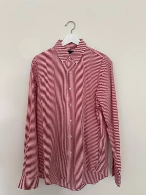 Polo Ralph Lauren skjorta - Säljer en röd- och vitrandig skjorta från Polo Ralph Lauren med klassisk knappkrage. Tidlös modell i hög kvalitet som passar både till vardag och mer uppklädda tillfällen. Använd ett fåtal gånger och är i nyskick. Nypris ca 1 200 kr.