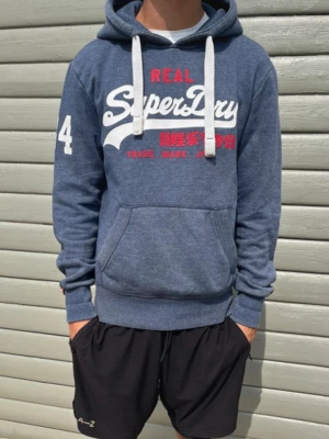 Blå Superdry hoodie med tryck - Superdry Hoodie - Mörkblå - Storlek S - Grymt skick! - Modellen på bilden är 181cm och väger 65kg. Tveka inte att skriva vid intresse!