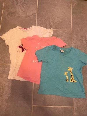 3 t-shirt för barn - Säljer en turkos t-shirt med korta ärmar och en söt ficka framtill där två gula giraffer sticker upp. T-shirten har en avslappnad passform och är gjord i mjuk bomull, perfekt för en chill och lekfull stil.