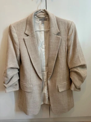 Beige kavaj med rynkad ärm - Stilren beige kavaj med klassisk krage och två fickor framtill. Kavajen har en strukturerad yta och är helfodrad med ljus insida. Ärmarna är lätt rynkade för en avslappnad look. Perfekt för dig som gillar en clean och tidlös stil.
