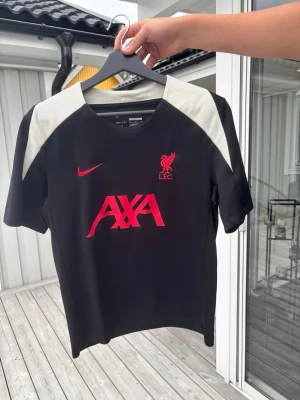 Svart Liverpool tränings t-shirt Nike - Snygg svart tränings t-shirt från Nike med Liverpool FC-logga och AXA sponsortryck i rött på bröstet. T-shirten är köpt på Anfield.