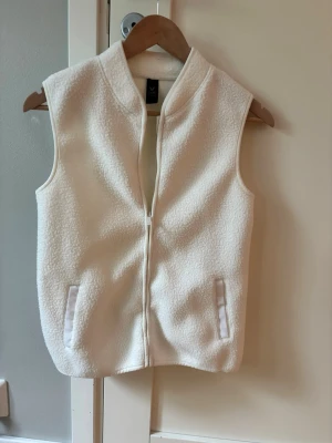 Vit pileväst med dragkedja - Snygg vit pileväst med dragkedja framtill och två fickor. Västen har en enkel och stilren design utan synligt märke, perfekt att slänga på sig över en hoodie eller tröja. Finns en lite röd fläck-se bild. Kan ev försvinna med fläckborttagningsmedel. Sparsamr använd väst. Fint skick i övrigt.