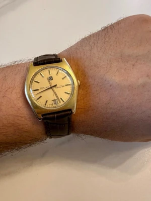 Vintage Certina Argonaut guldfärgad klocka med Morellato läderarmband  - Stilren Certina klocka med guldfärgat boett och ljus urtavla. Klockan har datumvisning och klassiska svarta index. Armbandet är brunt och gjort i läder, vilket ger en tidlös och elegant look. Fungerande, okänd service historik. Service behov kan förekomma 