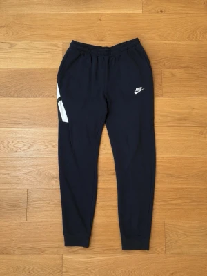 Mörkblå Nike mjukisbyxor - Säljer ett par mörkblå mjukisbyxor från Nike med vit logga på vänster lår och vita detaljer på sidan. Byxorna har resår i midjan och muddar vid bensluten. Perfekta för chill eller träning.
