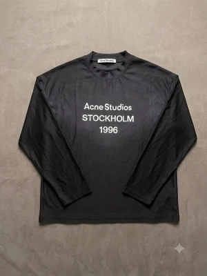Svart långärmad topp Acne Studios - Svart långärmad topp från Acne Studios med vit text 'Acne Studios STOCKHOLM 1996' på bröstet. Klassisk rund halsringning och rak passform. Perfekt för dig som gillar stilrena och trendiga plagg.