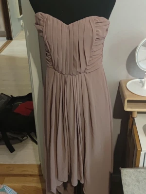 Gammelrosa axeöbandslös klänning stl 38 - Säljer en elegant gammelrosa klänning med axelbandslös design och draperad överdel. Kjolen är asymmetrisk och faller mjukt i flera lager. Perfekt för dig som vill ha en stilren och romantisk look.