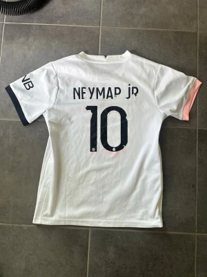PSG Neymar Jr matchtröja vit/rosa - Säljer en snygg PSG matchtröja i vitt med rosa och svart detalj, Neymar Jr och nummer 10 på ryggen. Tröjan har korta ärmar, rund hals och tryck på både fram- och baksida. Perfekt för dig som älskar fotboll och vill stötta Paris Saint-Germain.