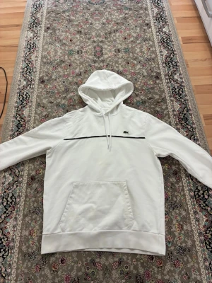 Vit hoodie med svart detalj från Lacoste - Säljer en stilren vit hoodie från Lacoste med svart linje över bröstet och klassisk krokodil-logga. Hoodien har huva med dragsko och en stor magficka. Perfekt för dig som gillar enkel men snygg design.