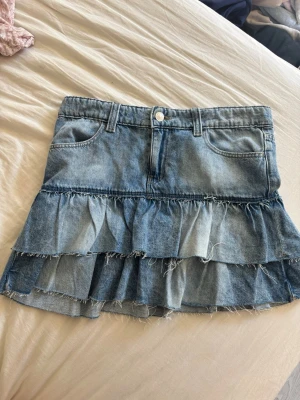 Blå jeanskjol med volanger - Säljer en jeanskjol i ljusblå denim med två lager volanger och råa kanter. Kjolen har klassiska fickor fram och bak samt hällor för skärp. Perfekt för dig som gillar en avslappnad men trendig stil.