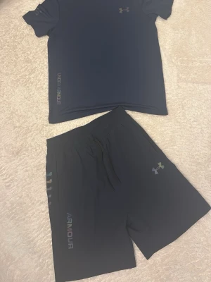 Mörk blå träningsset från Under Armour - Snyggt mörk blå träningsset från Under Armour med t-shirt och shorts. Båda plaggen har logga och text i regnbågsskimrande färg. Shortsen har dragsko i midjan och sidofickor. Perfekt för gymmet eller löprundan.