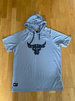 Ljusgrå hoodie med kort ärm och tryck - Ljusgrå hoodie med korta ärmar och svart tryck av en tjur på bröstet. Hoodien har svarta dragsnören och är tillverkad i ett tunt, mjukt material som känns sportigt. Perfekt för en avslappnad och cool stil.