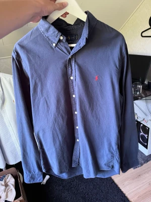 Marinblå skjorta med röd logga - Klassisk marinblå skjorta i bomull med knappar framtill och en broderad röd logga på bröstet. Skjortan har lång ärm och button-down krage, perfekt för en clean och stilren look.