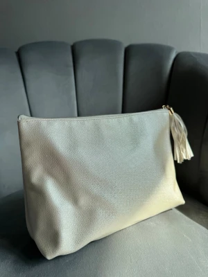 Beige smink väska med tofsdetalj - Stilren beige kuvertväska i ett mjukt, strukturerat material. Väskan har dragkedja upptill med en dekorativ tofs i matchande färg. Perfekt att bära i handen för en clean och modern look.