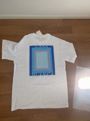 Vit t-shirt med blått tryck Casablanca - Vit t-shirt från Casablanca med ett stort grafiskt tryck i olika blå nyanser och texten 'CASA BLANCA' på ryggen. Mindre tryck framtill. Klassisk passform och rund hals. Perfekt för dig som gillar stilrena men ändå unika plagg.