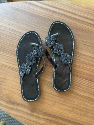 Svarta sandaler med pärlblommor - Säljer ett par svarta sandaler i läder med dekorativa pärlbroderade blommor på remmarna. Sandalerna har vita kontrastsömmar runt sulan och en klassisk tårem. Perfekta för dig som gillar unika detaljer och handgjorda inslag. Köpta i Kenya. Strl 39 men dom är som en 38