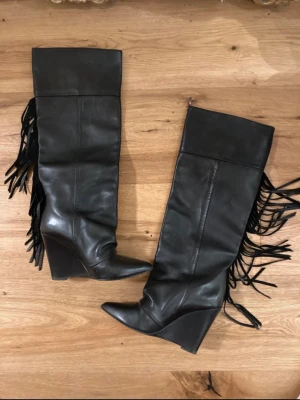 Svarta boots med kilklack och fransar - Snygga svarta stövlar i skinnimitation med hög kilklack och fransdetaljer längs sidan. Stövlarna har spetsig tå och når upp till knäna.  Storlek 36-37