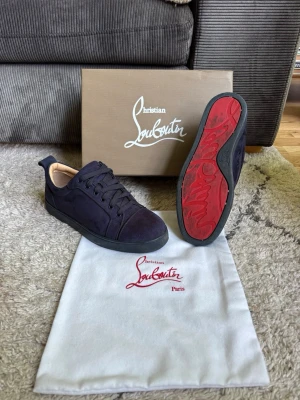 Mörkblå sneakers från Christian Louboutin - Snygga mörkblå sneakers i mocka från Christian Louboutin med klassisk röd sula och svarta skosnören. Lågt skaft och stilren design som passar perfekt till streetwear eller en mer uppklädd look. Kommer med originalkartong och dustbag.