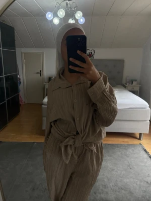 Beige ribbad loungewear-set - Säljer ett beige loungewear-set med ribbad struktur. Setet består av en långärmad skjorta med krage och knappar framtill samt matchande byxor med hög midja och knytband i midjan. Perfekt för chill dagar hemma eller när du vill vara bekväm men ändå se snygg ut. Finns några fläckar men går bort 