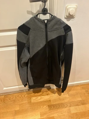 Svart och grå hoodie med dragkedja - Snygg hoodie i svart och grått med coola färgblock och hel dragkedja framtill. Tröjan har huva och är tillverkad i mjukt material, perfekt för en avslappnad streetstyle-look. Passar dig som gillar bekväma och stilrena plagg.