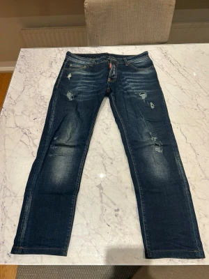 Dsquared 2 jeans - Storlek 42, italiensk storlek! Säljer ett par mörkblå jeans med raka ben och slitningar på flera ställen. Jeansen har klassisk femficksdesign, ljusa tvättade partier och detaljerade sömmar. Perfekta för en avslappnad och trendig look.