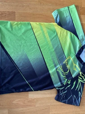 Grön sporttröja med vargmotiv - Säljer en sportig t-shirt i grönt och mörkblått med grafiska linjer och ett stiliserat vargmotiv på både fram- och baksida. Tillverkad i ett lätt och snabbtorkande material, perfekt för träning eller löpning. Tröjan har korta ärmar och rund hals.