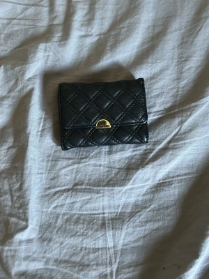 Svart quiltad plånbok i skinnimitation - Snygg svart plånbok i skinnimitation med quiltat mönster och guldfärgade detaljer. Framsidan har ett elegant spänne och baksidan har en praktisk ficka med dragkedja. Perfekt storlek för kort och kontanter.
