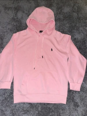 Ljusrosa hoodie med broderad logga - Säljer en ljusrosa hoodie med huva och dragsnören. Hoodien har en broderad liten logga på bröstet och en stor magficka framtill. Mjuk och skön bomullsblandning, perfekt för en chill look.