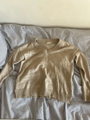 Beige stickad tröja med v-ringning - Säljer en beige stickad tröja med v-ringad hals och ribbade muddar vid ärmslut och nederkant. Tröjan har en avslappnad passform och långa ärmar, perfekt för lager på lager under kyligare dagar.