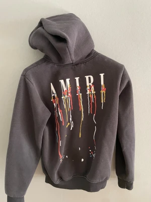 Svart hoodie från Amiri med tryck - Säljer en svart hoodie från Amiri med färgstarkt droppande tryck och stor logga på ryggen. Framsidan har en mindre version av trycket och känguruficka. Hoodien har ribbade muddar och är gjord i mjukt material. Perfekt för dig som gillar streetwear och unik design.