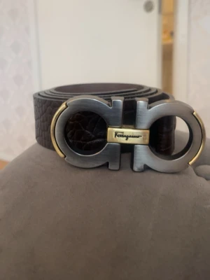 Brunt skärp med metallspänne Ferragamo - Snyggt brunt skärp från Ferragamo med ett ikoniskt metallspänne i silver och guld. Bältet har en präglad struktur som ger en lyxig känsla och passar perfekt till jeans eller kostymbyxor.