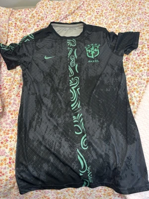 Svart Brasilien fotbollströja Nike - Snygg svart fotbollströja från Nike med Brasilien-logga och turkosa detaljer. Tröjan har korta ärmar, rund hals och ett unikt mönster i turkos som går vertikalt över hela tröjan. Perfekt för träning eller match.