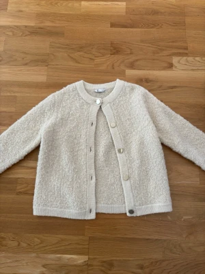 Krämvit kofta med rund hals - Mysig krämvit kofta med rund hals och stora silvriga knappar. Koftan har en fluffig, texturerad yta och ribbade muddar vid hals, ärmslut och nederkant. Perfekt för lager på lager och enkel att matcha med olika outfits.