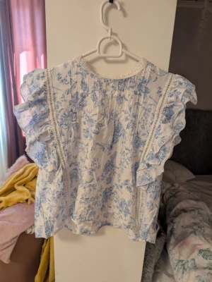 Blommig blå blus med volanger och spets - Söt blus i vitt med ljusblått blommönster och volanger längs ärmarna. Blusen har spetsdetaljer vid halsringningen och dekorativa sömmar framtill. Perfekt för en romantisk och somrig look.