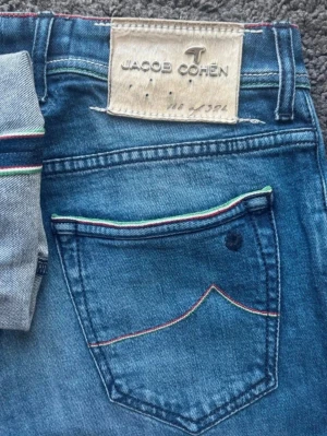 Blå jeans från Jacob Cohen - Säljer ett par blå jeans från Jacob Cohen med snygga detaljer i rött, vitt och grönt på bakfickan och sömmarna. Klassisk femficksmodell med patch i mocka baktill. Perfekta för dig som gillar stilrena men unika jeans.