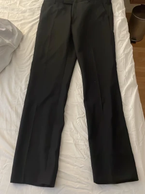 Svarta kostymbyxor med raka ben - Snygga svarta kostymbyxor med raka ben och klassisk passform. Byxorna har hysk- och hakstängning, dragkedja och knapp i midjan. Perfekta för en clean och stilren look. Materialet känns slätt och dressat.