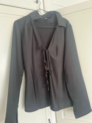 Svart blus med knytning i satinlook - Stilren svart blus i glansigt satinliknande material med djup v-ringning och knytband framtill. Långa ärmar och klassisk krage ger en elegant touch. Perfekt att styla med jeans eller kjol för en trendig look.