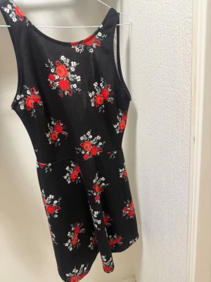Svart blommig kortklänning - Supersöt svart kortklänning med röda och vita blommor. Klänningen är ärmlös och har en v-ringad rygg som ger en snygg detalj. Perfekt för varma dagar eller när du vill känna dig extra fin. Materialet känns mjukt och följsamt.