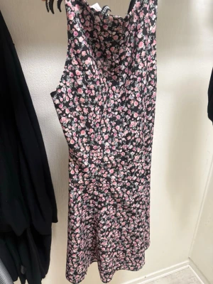 Svart blommig kortklänning - Söt svart kortklänning med rosa och vita blommor. Klänningen är ärmlös och har en rund halsringning. Perfekt för sommarens varma dagar och har en lätt utsvängd passform. Materialet känns mjukt och följsamt.