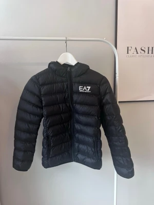 Svart dunjacka från EA7 Emporio Armani - Snygg svart dunjacka från EA7 Emporio Armani med huva och dragkedja framtill. Jackan har logga på bröstet och är i super bra skick!! Endast använd ett fåtal gånger och är istortsätt som ny! Storlek 12 år (150cm) NYPRIS = 1799