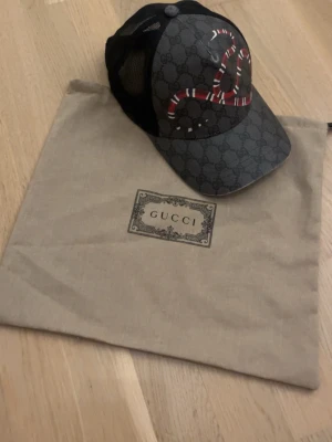 Gucci keps med orm och GG-mönster - Snygg keps från Gucci i mörkgrått med klassiskt GG-mönster och en rödvit orm broderad framtill. Baksidan har svart mesh för extra ventilation och justerbar rem. Perfekt accessoar för dig som vill sticka ut med lyxig streetstyle.