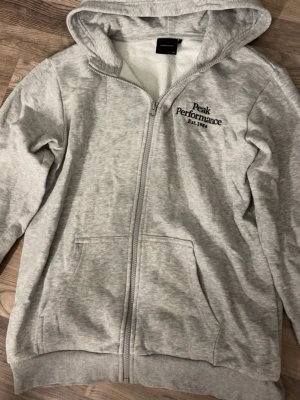 Grå zip hoodie från Peak Performance - Snygg ljusgrå hoodie från Peak Performance med dragkedja och broderad logga på bröstet. Mjuk insida i fleece, två fickor fram och klassisk huva. Perfekt för chill dagar eller när du vill ha en enkel men stilren look.