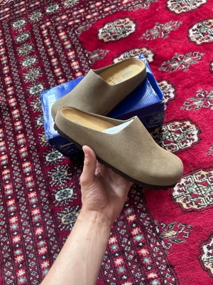 Beige Birkenstock tofflor i mocka - Säljer ett par snygga beige tofflor från Birkenstock i mjuk mocka. De har en klassisk slip-in modell med rund tå och bekväm innersula. Perfekta för att chilla hemma eller snabbt dra på sig när man ska ut. Kommer i originalkartong.