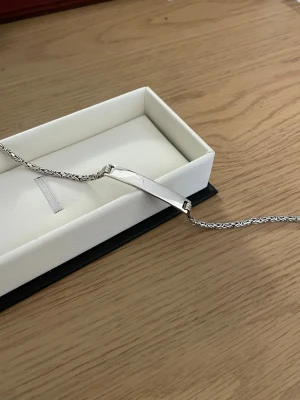 Silverfärgat kedjearmband med platta - Stilrent armband i silverfärg med en snygg, flätad kedja och en slät rektangulär platta i mitten. Perfekt för att gravera in ett namn eller budskap. Armbandet har en klassisk och tidlös design som passar till många olika stilar.