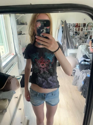 Svart t-shirt med färgglatt tryck - Säljer en svart t-shirt med färgglatt grafiskt tryck i blått, rött och lila på framsidan. T-shirten har rund hals och korta ärmar. Perfekt för dig som gillar en edgy och unik stil.